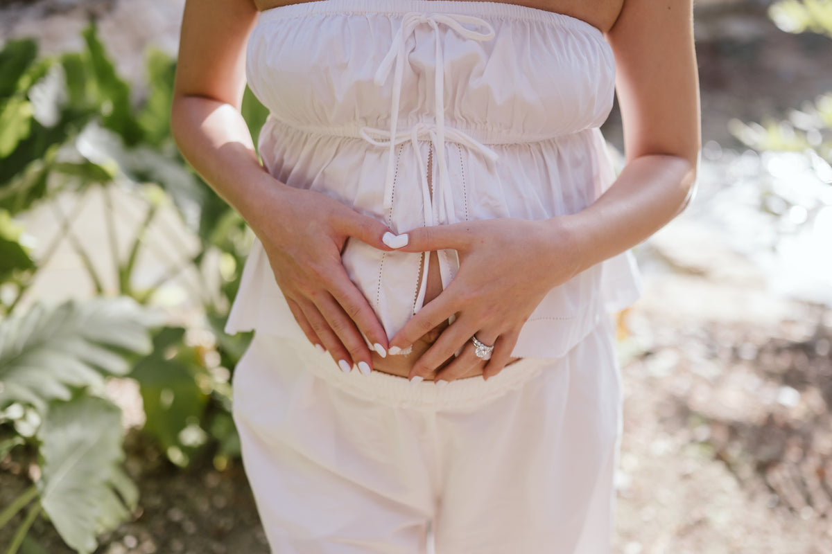 Expecting mother in white makes a heart with her hands over her bump in a tropical garden