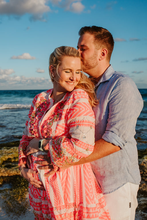 Man kisses his pregnant partner's head as they cradle her bump on a rocky shore at sunset