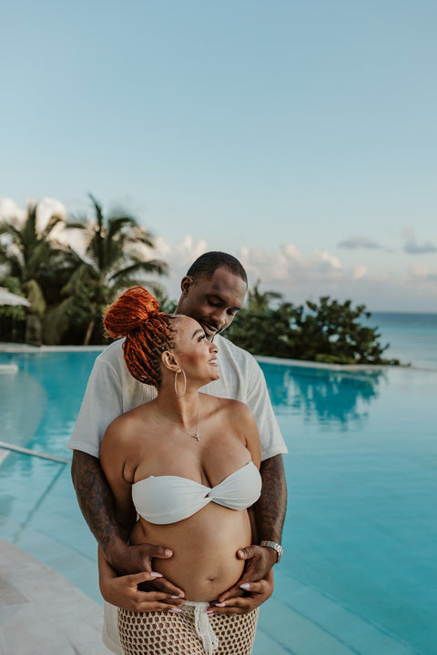 Partner hugs a mother-to-be from behind beside an infinity pool during a resort maternity photoshoot