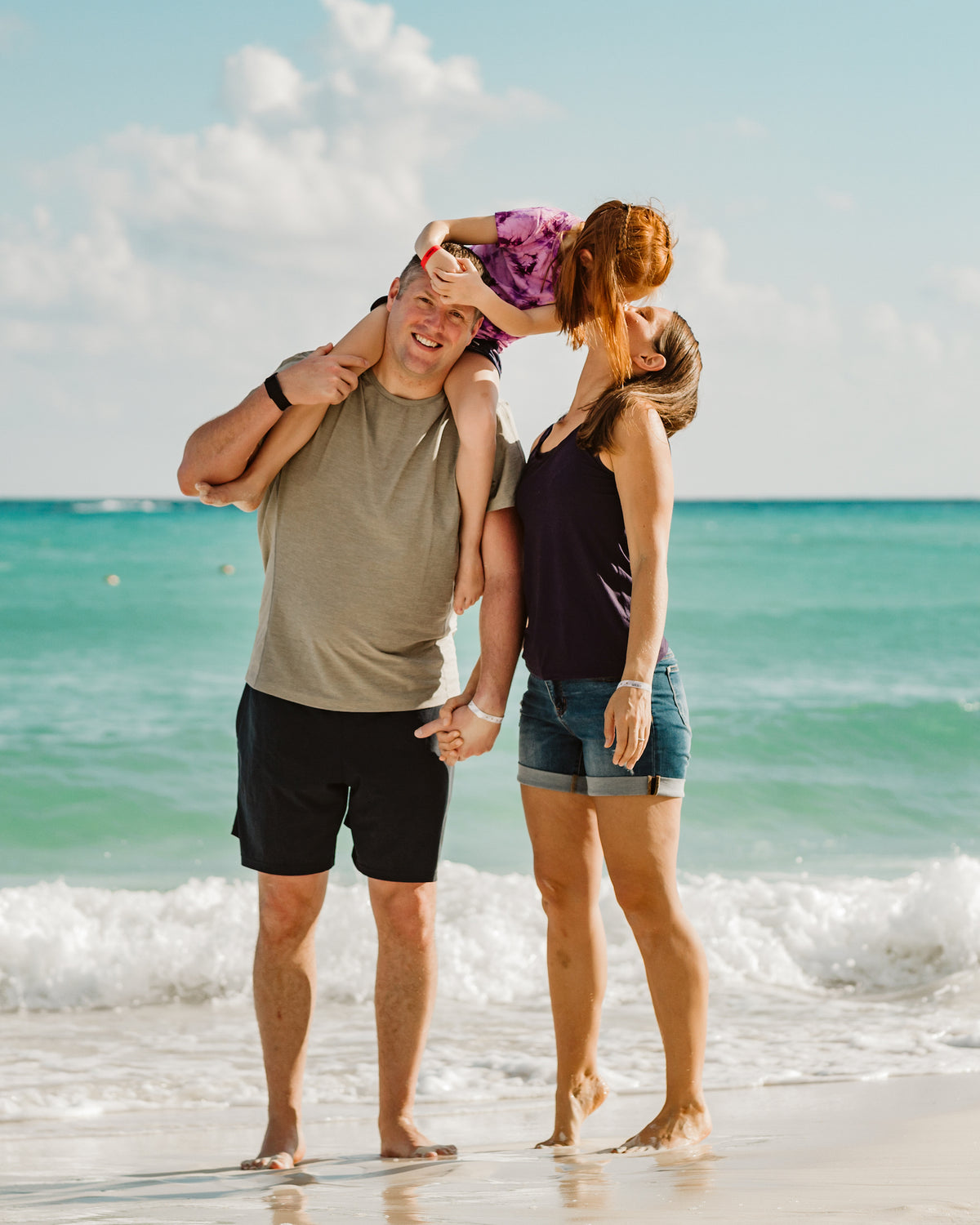 Daughter on her dad's shoulders kisses her mom on a turquoise Mexican beach