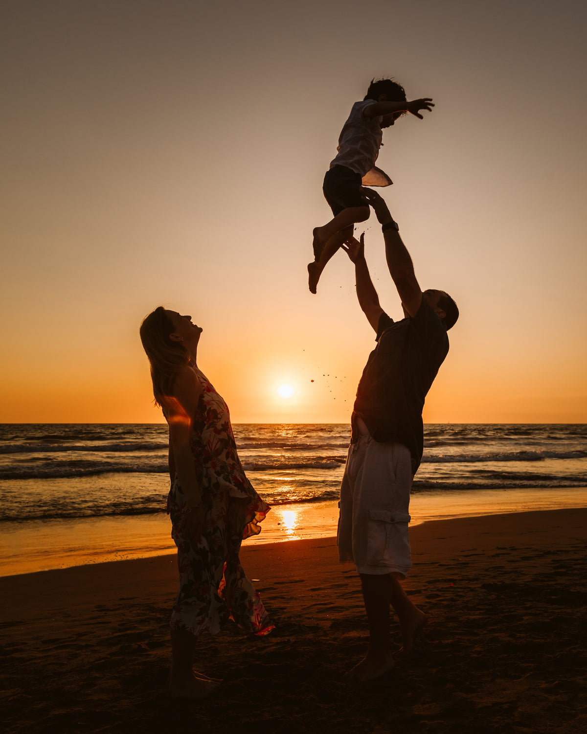Silhouette of a dad tossing his child into the air at sunset as mom watches on the beach