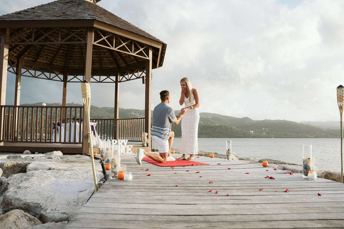 Man kneels to propose on a rose-petal dock beside a gazebo and tiki torches in Montego Bay