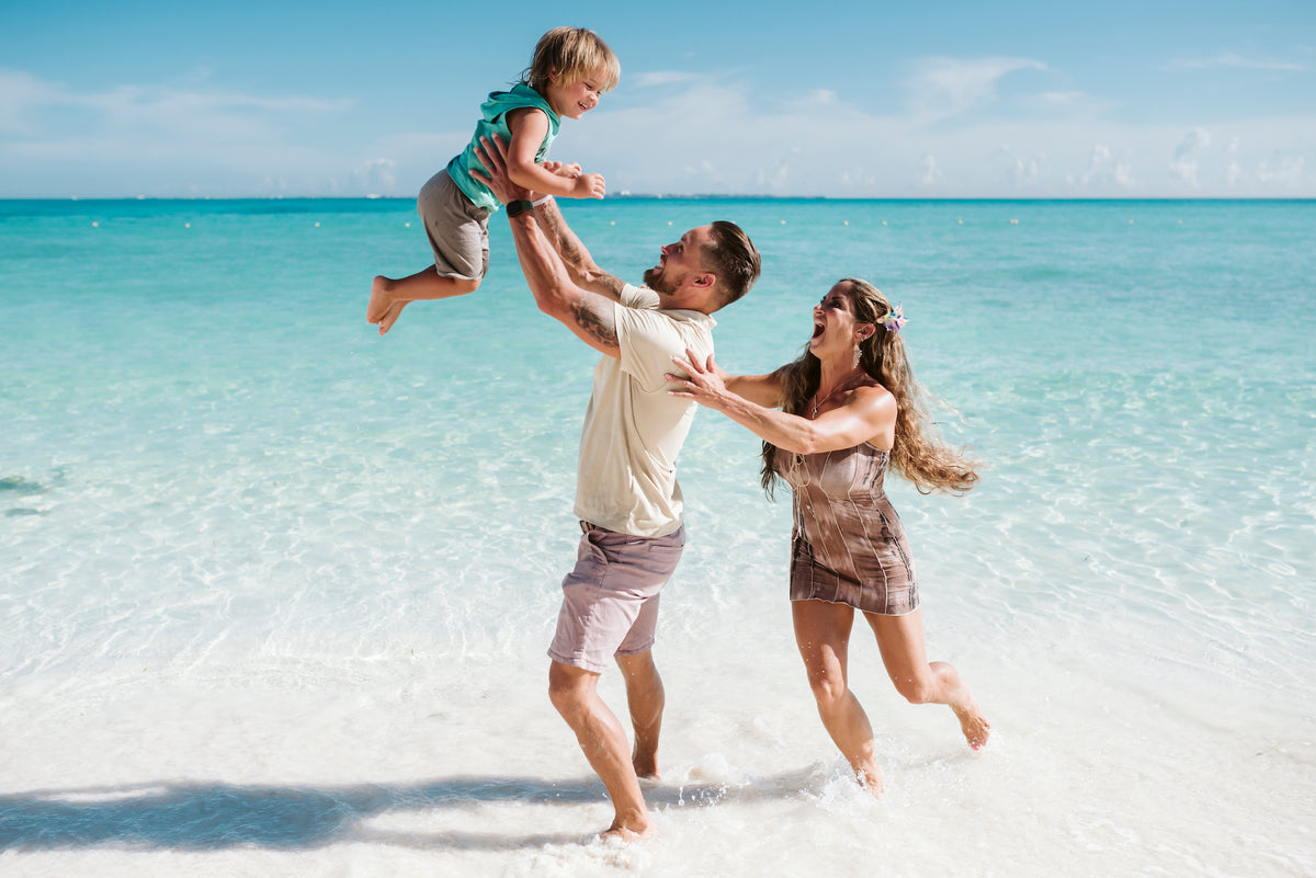 Father tosses his young son in the air as mom laughs in turquoise shallow water in Cancun