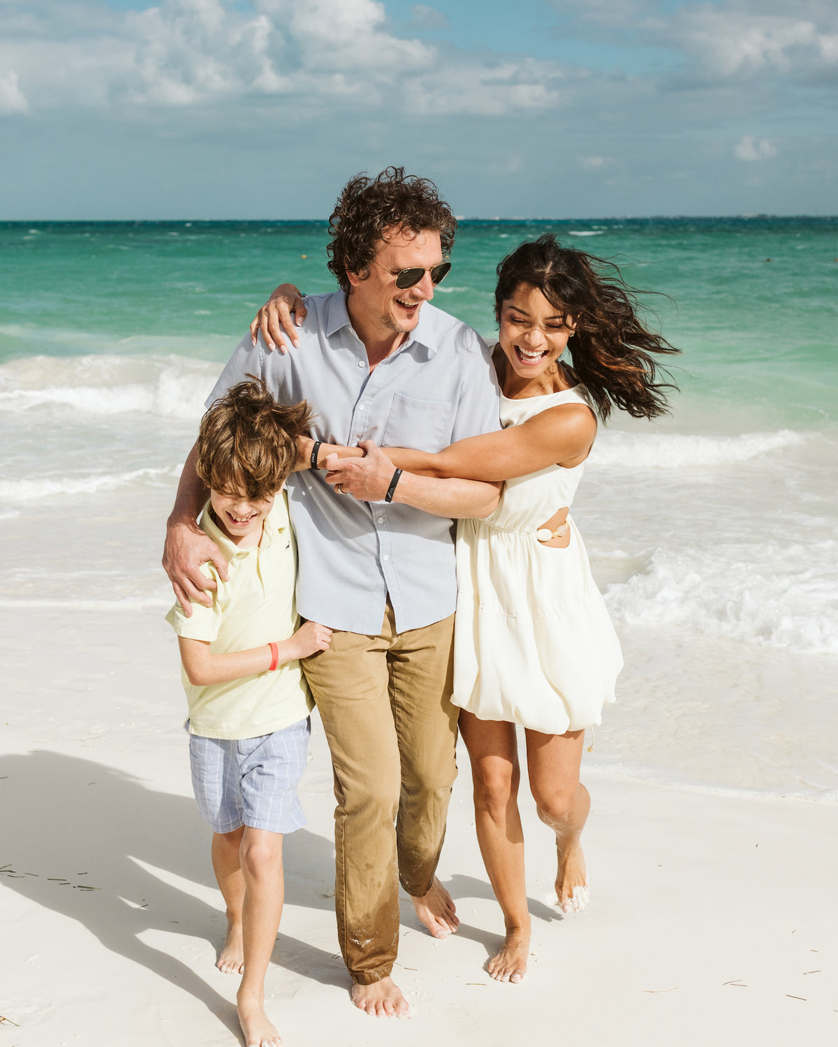 Laughing family of three walks arm in arm along a white sand beach during a family photoshoot in Mexico