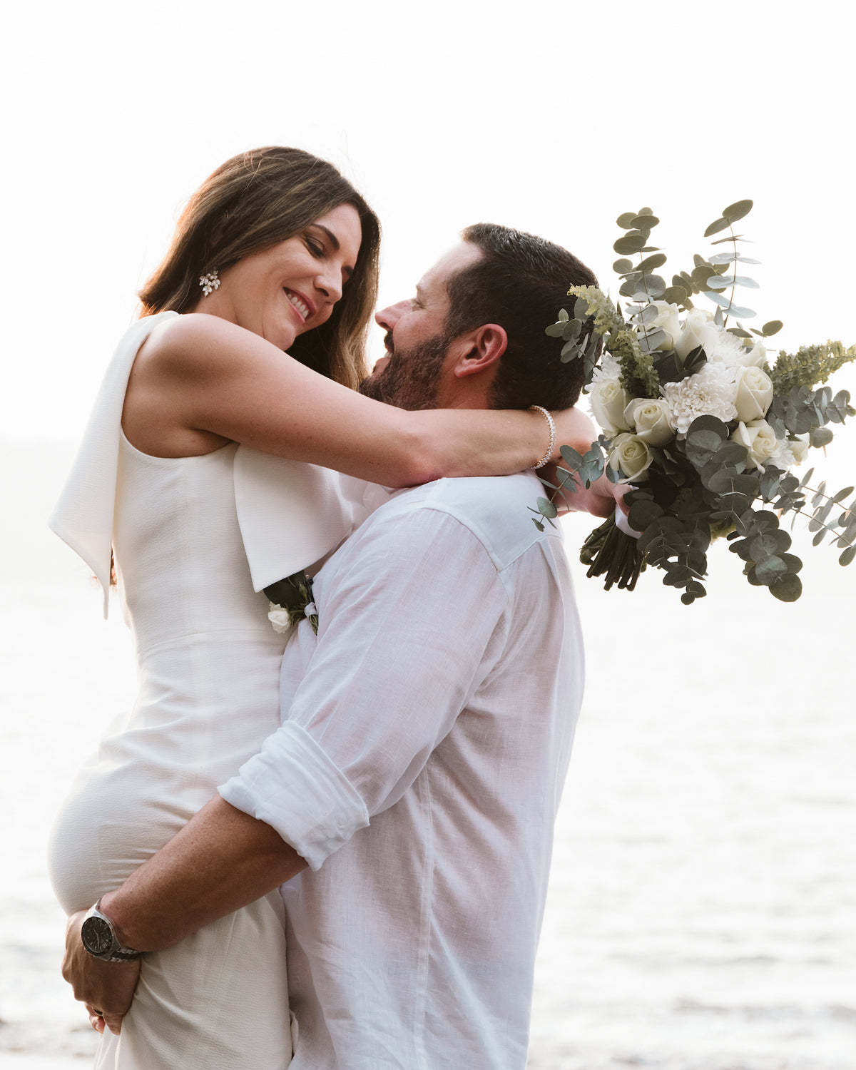 Bride wraps her arms around the groom as he lifts her by the sea on their wedding day