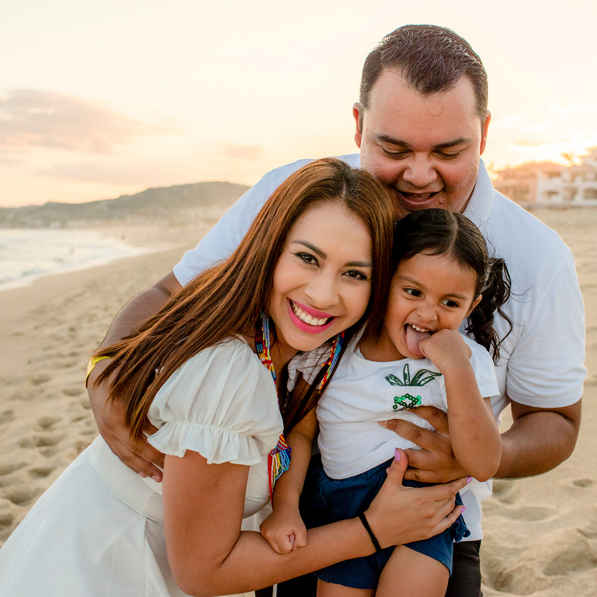 Parents hug their laughing young daughter on the beach at sunset at Vidanta Los Cabos