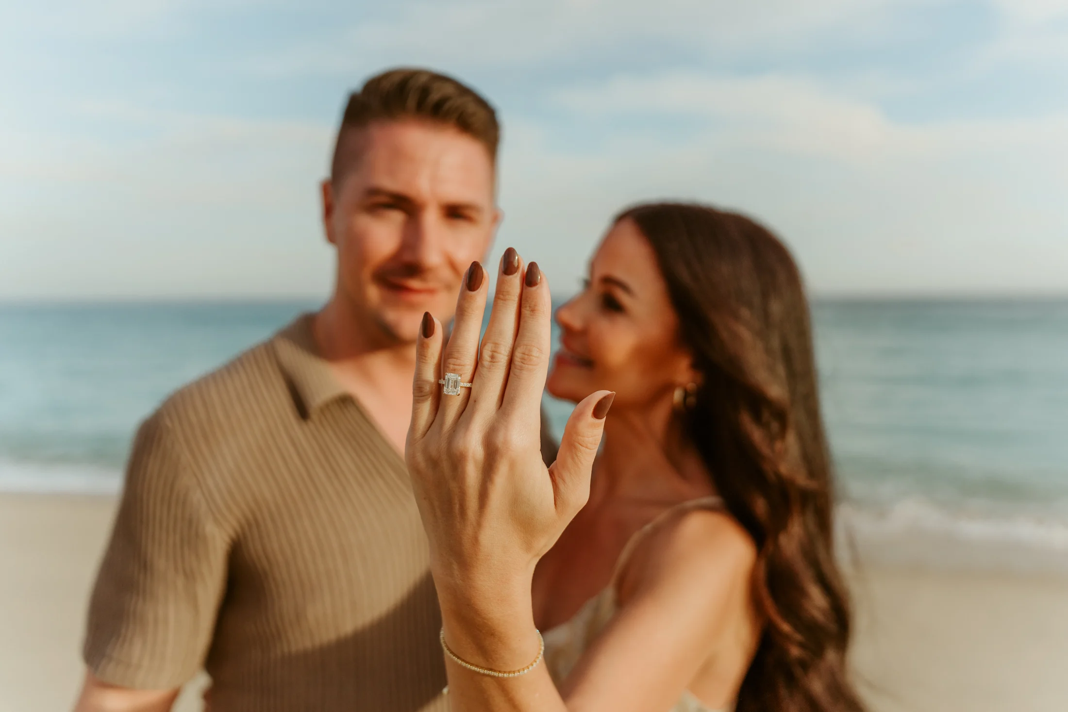 A newly engaged woman shows her ring beside her fiancé on the beach